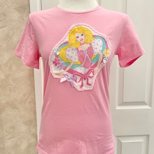 May include: Pink t-shirt featuring a colourful appliqu&eacute; of a Barbie doll wearing a pink dress and a crown. The appliqu&eacute; has a white lace border and pink bows.