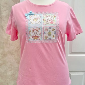 K&ouml;nnte beinhalten: Ein hellrosa T-Shirt mit einem quadratischen Aufn&auml;her mit Cartoon-Hasen und anderen niedlichen Illustrationen. Eine hellblaue Schleife ist am Shirt befestigt. Das Shirt wird auf einer Schaufensterpuppe pr&auml;sentiert.