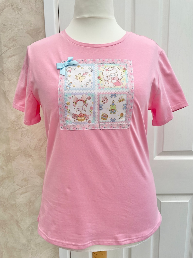 K&ouml;nnte beinhalten: Ein hellrosa T-Shirt mit einem quadratischen Aufn&auml;her mit Cartoon-Hasen und anderen niedlichen Illustrationen. Eine hellblaue Schleife ist am Shirt befestigt. Das Shirt wird auf einer Schaufensterpuppe pr&auml;sentiert.