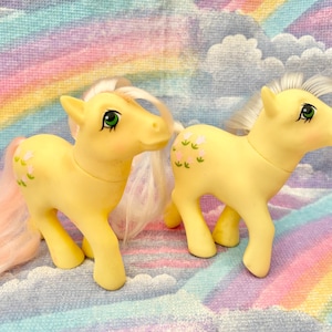 May include: Two vintage yellow toy ponies with pink and white manes and tails. Each pony has green eyes and a floral design on its flank. The ponies are standing on a pastel rainbow background.
