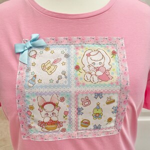 K&ouml;nnte beinhalten: Hellrosa T-Shirt mit einem quadratischen Patch mit Cartoon-Figuren und floralen Mustern. Eine hellblaue Schleife ist angebracht. Der Patch hat einen rosa-wei&szlig;en Blumenrand. Das T-Shirt besteht aus einem weichen Material.