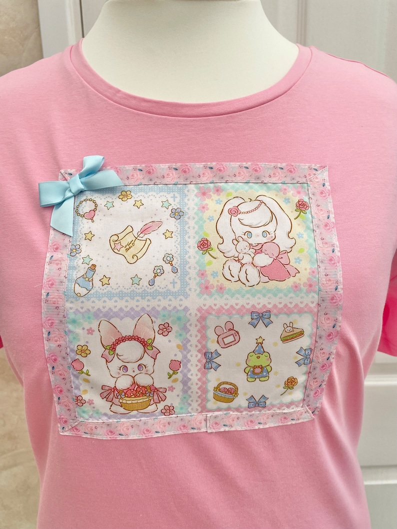 K&ouml;nnte beinhalten: Hellrosa T-Shirt mit einem quadratischen Patch mit Cartoon-Figuren und floralen Mustern. Eine hellblaue Schleife ist angebracht. Der Patch hat einen rosa-wei&szlig;en Blumenrand. Das T-Shirt besteht aus einem weichen Material.