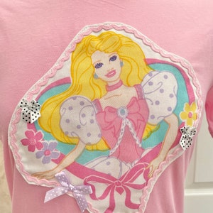 May include: Pink t-shirt with a large, heart-shaped appliqu&eacute; featuring a vintage Barbie doll illustration. The appliqu&eacute; has a pink, white and blue colour scheme and is surrounded by a white lace trim. The t-shirt has a pink bow with white polka dots on the bottom left corner.
