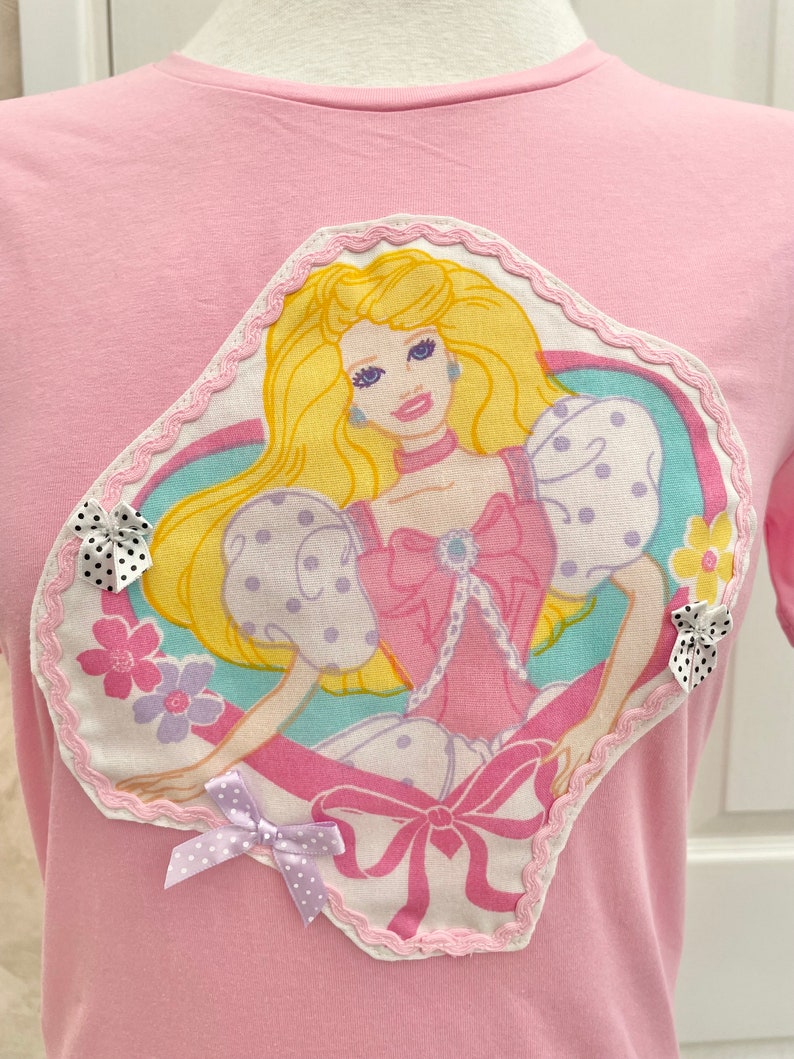 May include: Pink t-shirt with a large, heart-shaped appliqu&eacute; featuring a vintage Barbie doll illustration. The appliqu&eacute; has a pink, white and blue colour scheme and is surrounded by a white lace trim. The t-shirt has a pink bow with white polka dots on the bottom left corner.