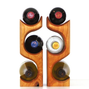 May include: Two wooden wine racks, each holding two wine bottles. The racks are made of light-colored wood with a curved design. The wine bottles are various colors, including red, black, and clear.