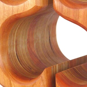May include: Wooden wine rack with a layered design. The rack is made of light and dark brown wood, with curved openings for bottles. The design features a series of curved, layered wooden elements.