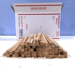 May include: A pile of small, square wooden dowels in varying shades of brown. The dowels are stacked in a neat arrangement on a white surface. A USPS Priority Mail box is in the background, with text that reads "SMALL FLAT RATE BOX".