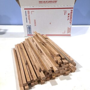 May include: A pile of square wooden dowels with a natural wood grain finish. A white USPS Priority Mail box is in the background. The box has red and black text, including "Priority Mail" and "Small Flat Rate Box."