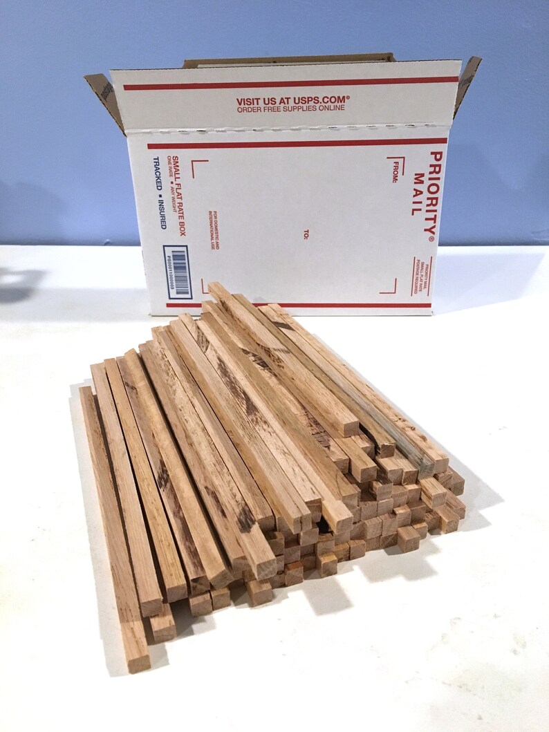 May include: A pile of square wooden dowels with a natural wood grain finish. A white USPS Priority Mail box is in the background. The box has red and black text, including "Priority Mail" and "Small Flat Rate Box."