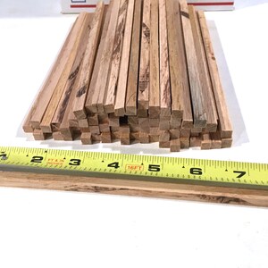 May include: A collection of small, square wooden dowels, likely for crafting or model building. The dowels are a natural wood color with visible grain. A yellow tape measure shows the dowels are approximately 1/4 inch thick.