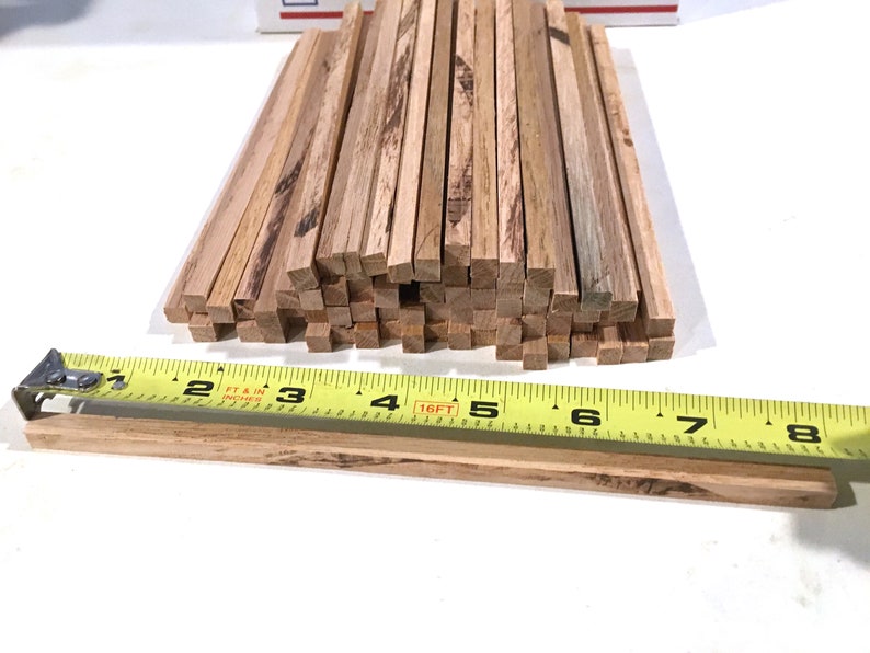 May include: A collection of small, square wooden dowels, likely for crafting or model building. The dowels are a natural wood color with visible grain. A yellow tape measure shows the dowels are approximately 1/4 inch thick.