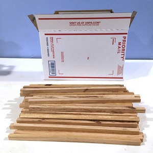 May include: A stack of unfinished wooden strips next to a white Priority Mail box. The box is open, displaying the text "VISIT US AT USPS.COM ORDER FREE SUPPLIES ONLINE" and "PRIORITY MAIL". The wooden strips are light brown.