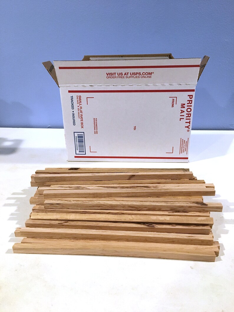 May include: A stack of unfinished wooden strips next to a white Priority Mail box. The box is open, displaying the text "VISIT US AT USPS.COM ORDER FREE SUPPLIES ONLINE" and "PRIORITY MAIL". The wooden strips are light brown.