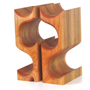 May include: A wooden wine rack with a unique, curved design. The rack is made of light brown wood and features openings for holding wine bottles. The design is modern and sculptural, suitable for home decor.