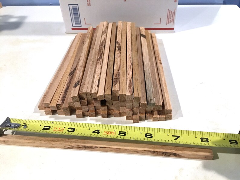 May include: A collection of small, square wooden dowels, approximately 9 inches long, with natural wood grain. A yellow measuring tape is placed next to the dowels for scale. The dowels are stacked in a pile, with some lying flat.