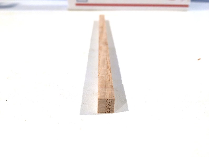 May include: A rectangular piece of light brown wood, possibly a small beam or strip, is centered on a white surface. The wood has a visible grain and a square end with small holes. The wood is resting on a thin, translucent white material.