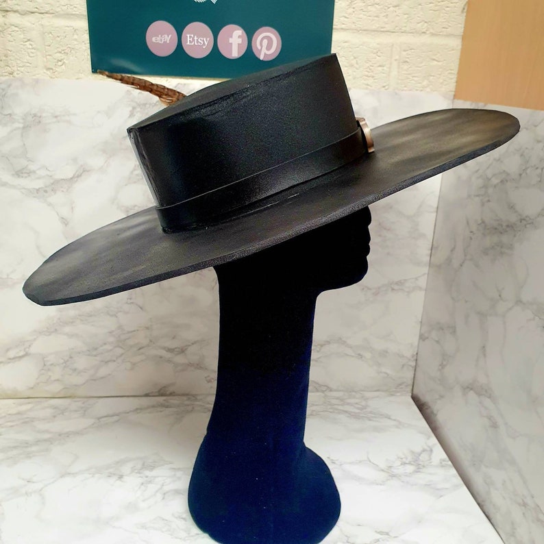Puede incluir: Un sombrero negro de ala ancha con una corona alta y plana. El sombrero tiene una banda negra alrededor de la corona y una hebilla plateada en el lateral.
