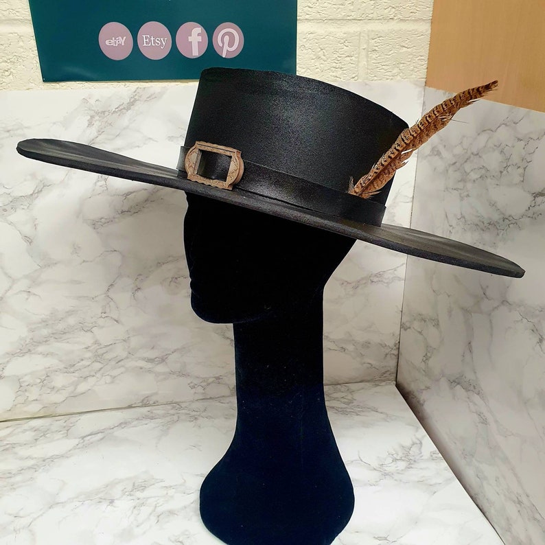 Puede incluir: Un sombrero negro de ala ancha con una pluma marr&oacute;n y una hebilla dorada. El sombrero est&aacute; en una cabeza de maniqu&iacute; negra.