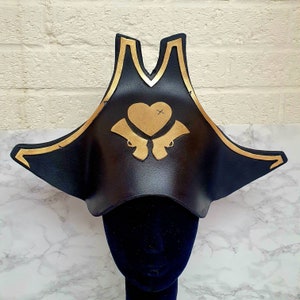 Puede incluir: Un sombrero de pirata negro con ribete dorado y un corazón dorado con pistolas cruzadas.