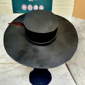 Puede incluir: Un sombrero negro de ala ancha con una corona alta y cil&iacute;ndrica. El sombrero tiene un adorno de plumas y se muestra en un soporte negro.