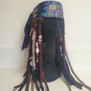 May include: A black mannequin head with long brown, black, and gray dreadlocks. The mannequin is wearing a blue and pink paisley bandana. The dreadlocks are decorated with silver beads and a silver cross charm.