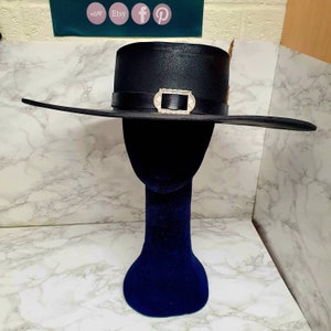 Puede incluir: Un sombrero negro de ala ancha con una hebilla plateada y un adorno de pluma. El sombrero est&aacute; en una cabeza de maniqu&iacute; azul.
