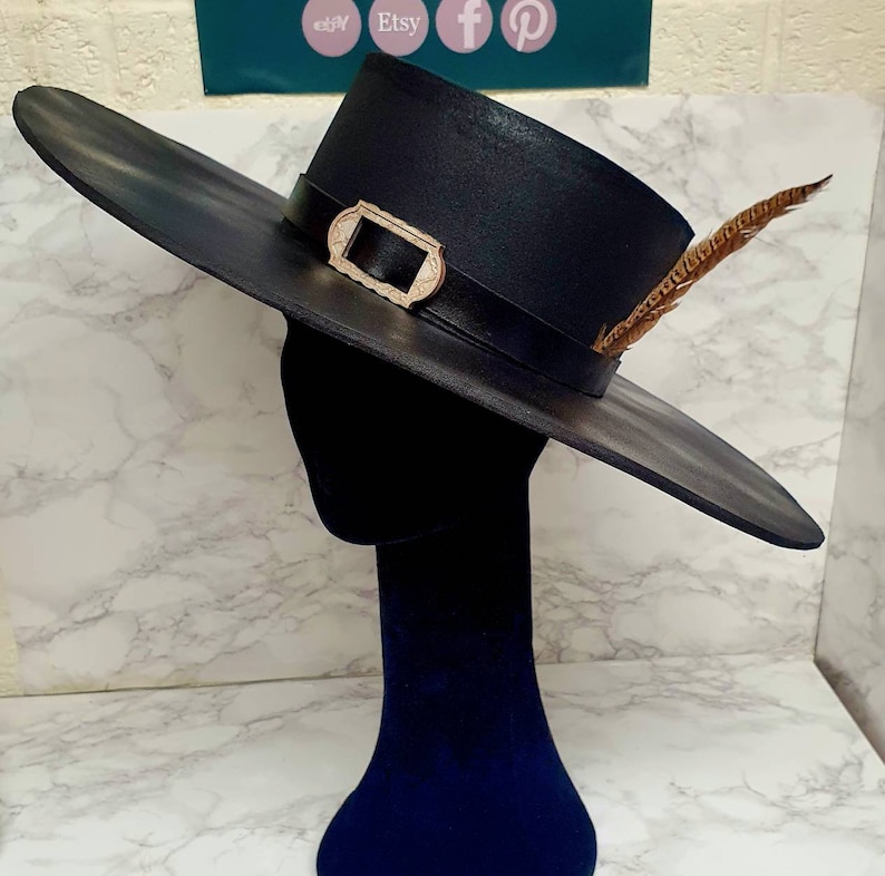 Puede incluir: Un sombrero negro de ala ancha con una hebilla dorada y un adorno de pluma marr&oacute;n. El sombrero est&aacute; hecho de un material liso y texturizado y tiene un ala ligeramente curvada.
