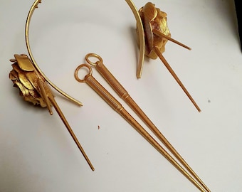 Puede incluir: Un conjunto de accesorios para el cabello de color dorado, que incluye una diadema con dientes, dos palillos para el pelo y dos piezas florales decorativas. El conjunto está hecho de un material metálico y está diseñado para ocasiones formales.
