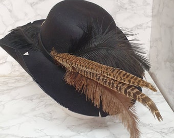 Puede incluir: Un sombrero negro con ala ancha, adornado con plumas oscuras y plumas marrones y beige estampadas. El sombrero tiene una copa redonda y una banda decorativa. Adecuado para disfraces o eventos formales.