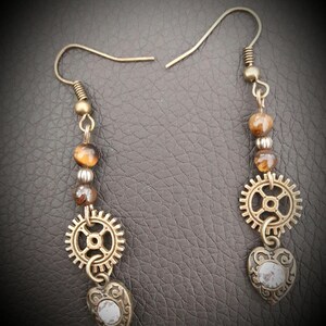 May include: A pair of gold-toned earrings with tiger's eye beads, a gear charm, and a heart charm with a clear crystal.