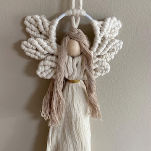 May include: A white macrame angel ornament with a wooden head and brown hair. The angel has wings and a long, flowing dress.