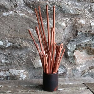 May include: A collection of wooden knitting needles in a black ceramic container. The needles are arranged in a radial pattern, with the tips pointing upwards.