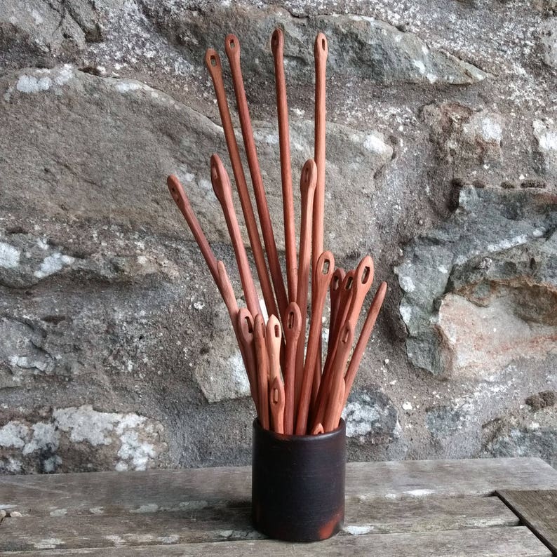 May include: A collection of wooden knitting needles in a black ceramic container. The needles are arranged in a radial pattern, with the tips pointing upwards.