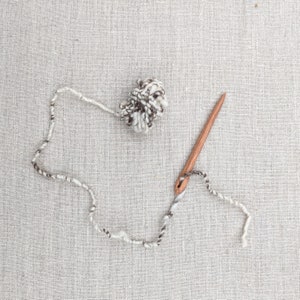 May include: A wooden needle with a white and brown yarn threaded through it, and a ball of white and brown yarn on a linen background.