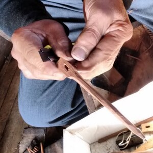 May include: A person's hands are shown holding a wooden dowel with a small metal piece attached to it. The dowel is being worked on with a tool, possibly a chisel. The dowel is light brown in color and has a smooth surface. The metal piece is dark gray and has a sharp edge.