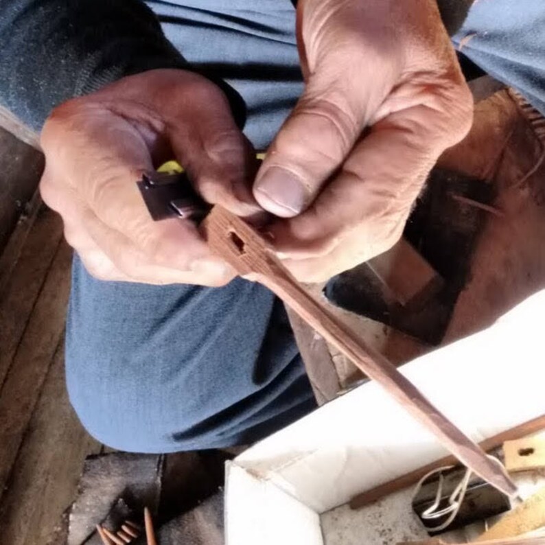 May include: A person's hands are shown holding a wooden dowel with a small metal piece attached to it. The dowel is being worked on with a tool, possibly a chisel. The dowel is light brown in color and has a smooth surface. The metal piece is dark gray and has a sharp edge.