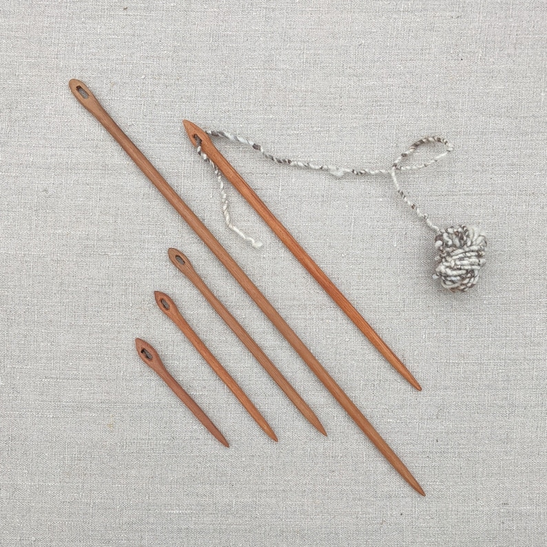 May include: Five wooden knitting needles of varying lengths, with one needle attached to a ball of white and gray yarn.