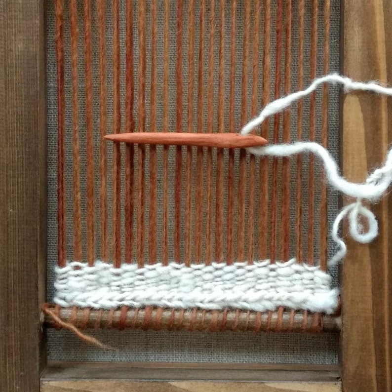 May include: A close-up of a wooden loom with a white yarn being woven into a brown fabric. A wooden shuttle is used to weave the yarn.