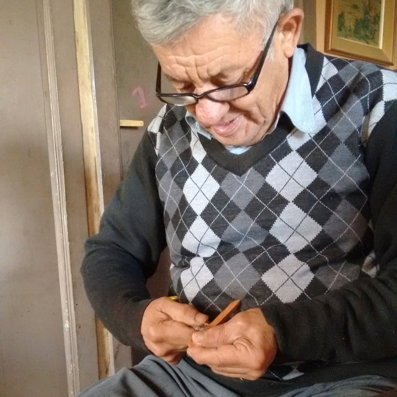 May include: A man wearing glasses and a black and grey sweater is sitting and working on a craft project. He is using a small tool to work on a piece of material.