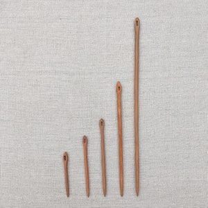 May include: Five wooden knitting needles of varying lengths, arranged in a graduated size order from smallest to largest. The needles are all brown and have a pointed tip and a small eye at the top.