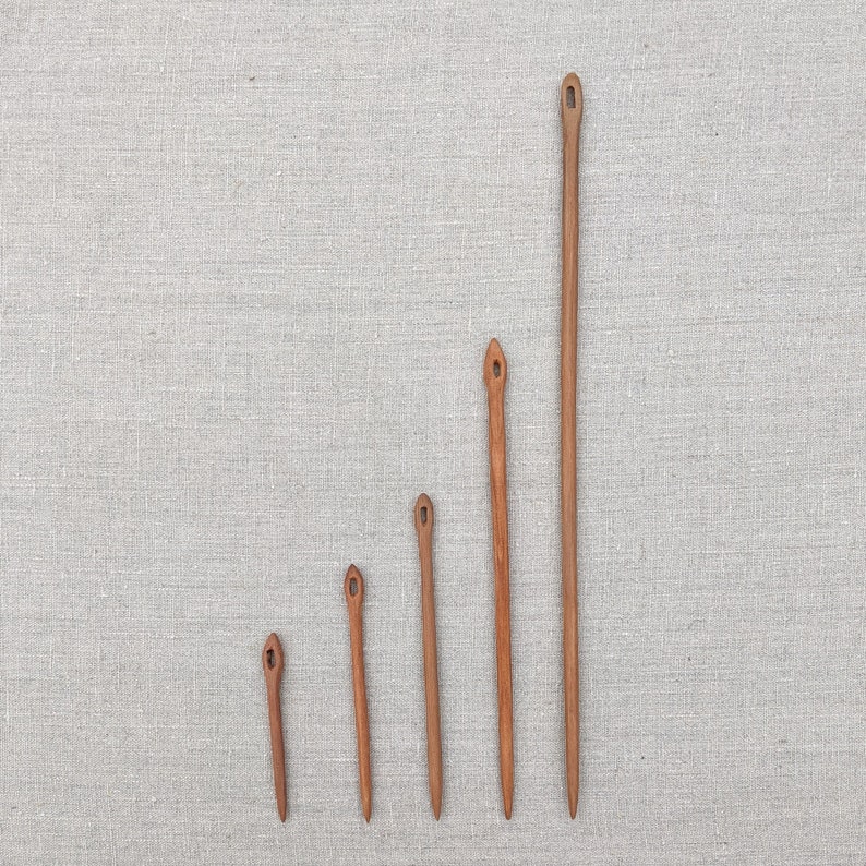 May include: Five wooden knitting needles of varying lengths, arranged in a graduated size order from smallest to largest. The needles are all brown and have a pointed tip and a small eye at the top.