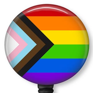 May include: A round, glossy button featuring the Pride flag with a rainbow of colors and the Progress Pride flag with pink, light blue, brown, black, and white. The button has a black backing.