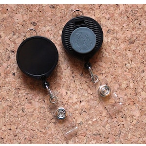 May include: Two black retractable badge holders with clear plastic straps. The badge holders have a round shape and a silver clip. One badge holder has a gray warning label that reads "Caution - Retractible Device".