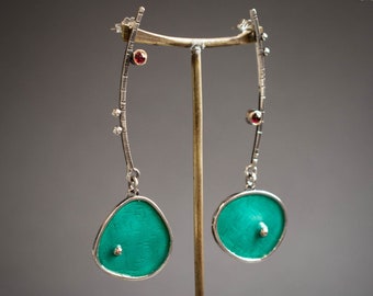 May include: A pair of silver earrings with a turquoise green oval pendant and red gemstones. The earrings are hanging from a stand.