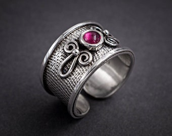 May include: A silver ring with a textured band and a pink gemstone set in a silver bezel. The ring has a decorative design on the band.