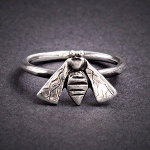 May include: A silver ring with a detailed bee design. The bee's wings are textured and the body is striped.