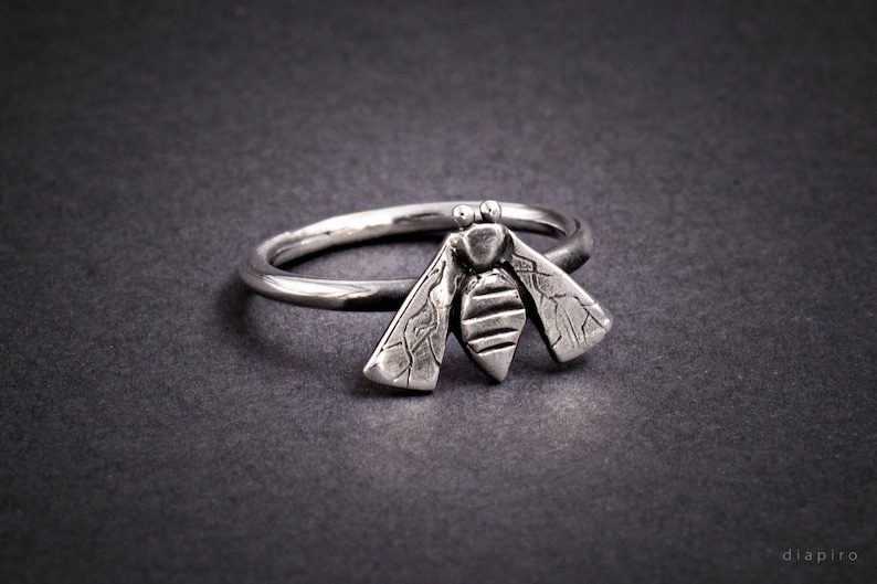 May include: A silver ring with a textured bee design. The bee has wings and a segmented body.