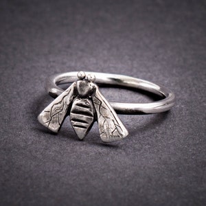 May include: A silver ring with a detailed bee design. The bee is textured and has wings that are spread out.