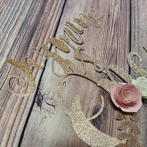May include: Gold glitter cake topper with the name "Jacqueline" and a floral design with pink and white paper flowers.
