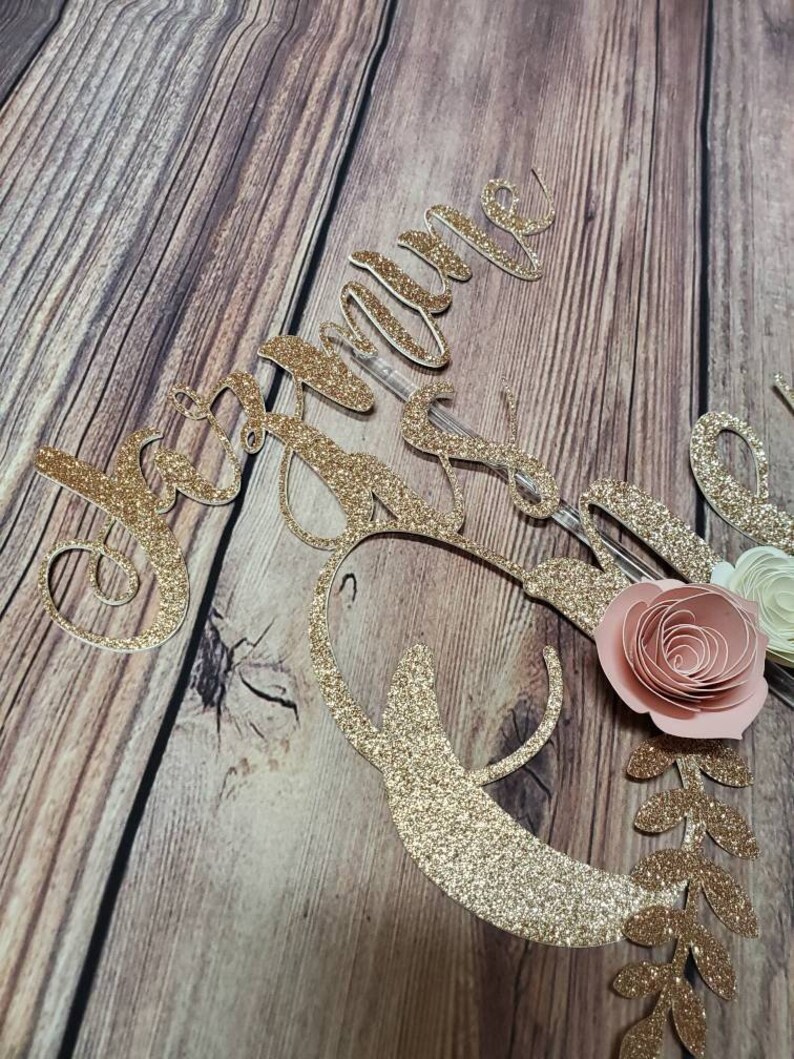 May include: Gold glitter cake topper with the name "Jacqueline" and a floral design with pink and white paper flowers.